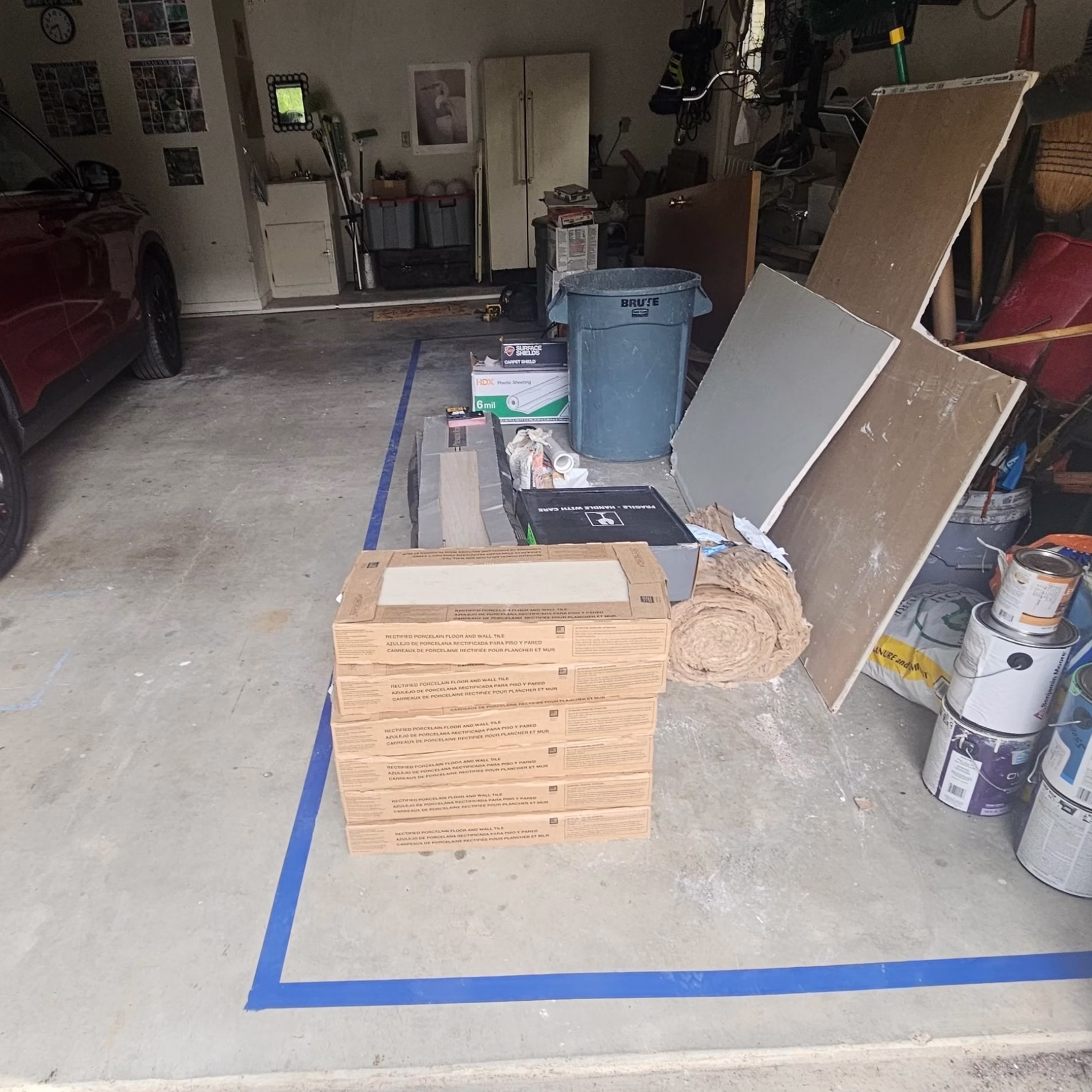 Remodeling materials and supplies organized inside a designated area marked with blue painter's tape on a residential garage floor.