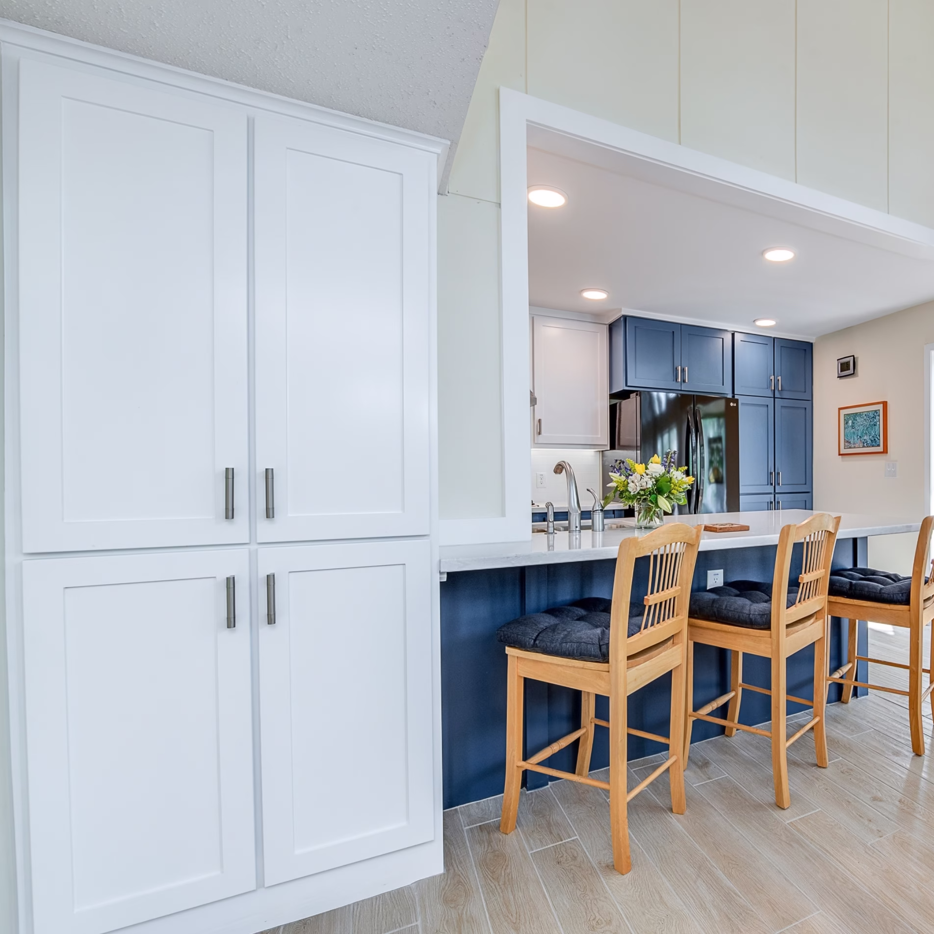 New white storage cabinets beside the enlarged kitchen opening and blue bar cabinetry with seating.