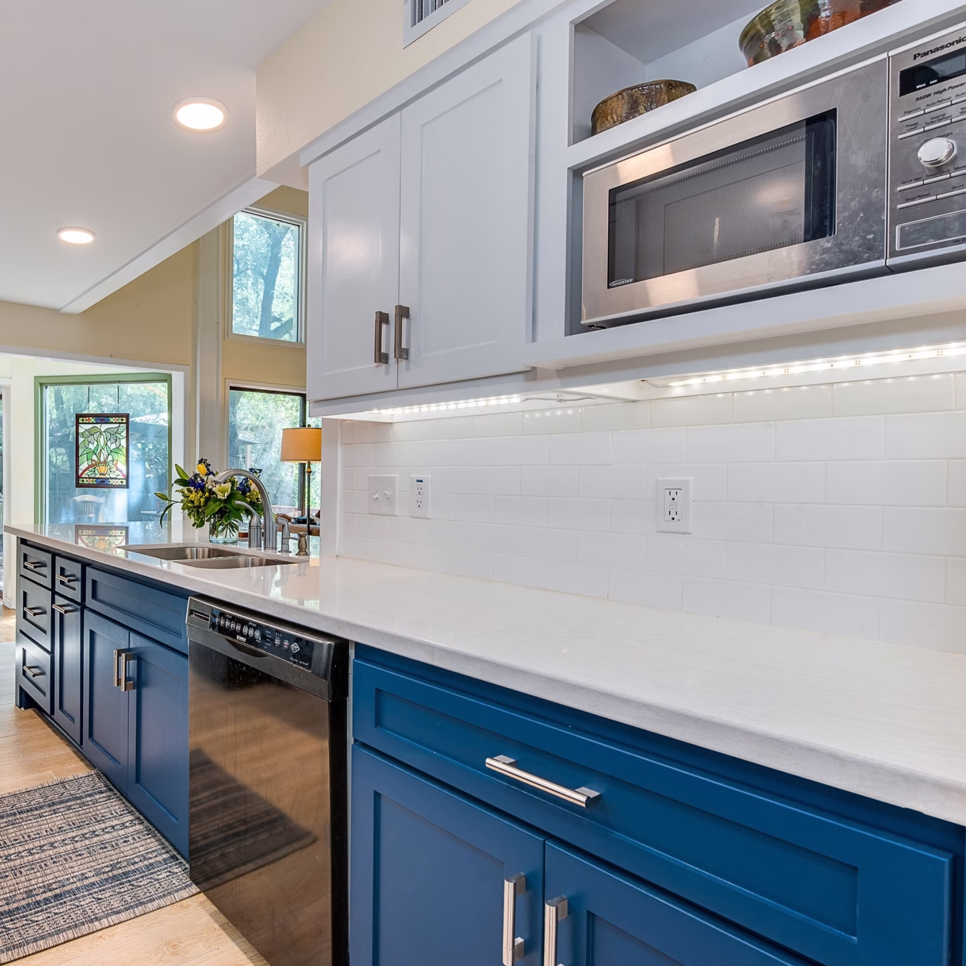 Remodeled sink side of the kitchen with blue base cabinets, white upper cabinets, white countertop, tile backsplash, and built-in microwave.