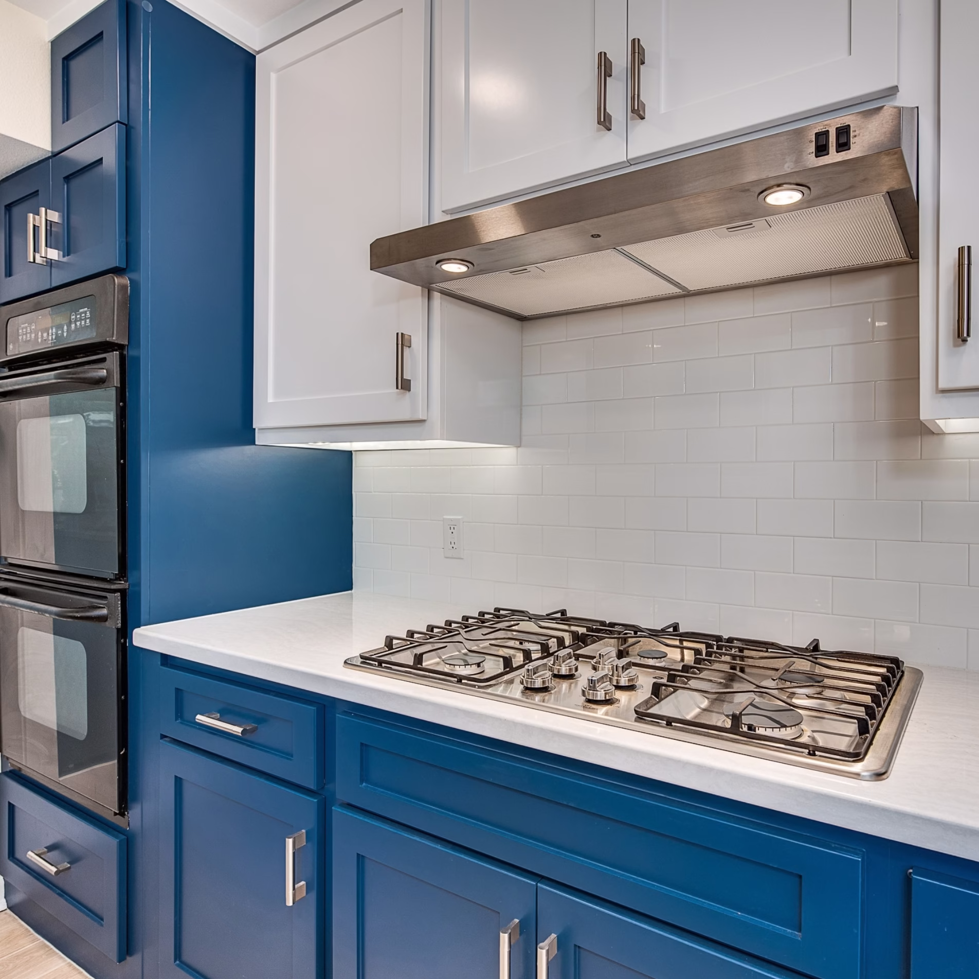 Remodeled cooking area with blue base cabinets, white upper cabinets, gas cooktop, stainless steel vent hood, and white tile backsplash.