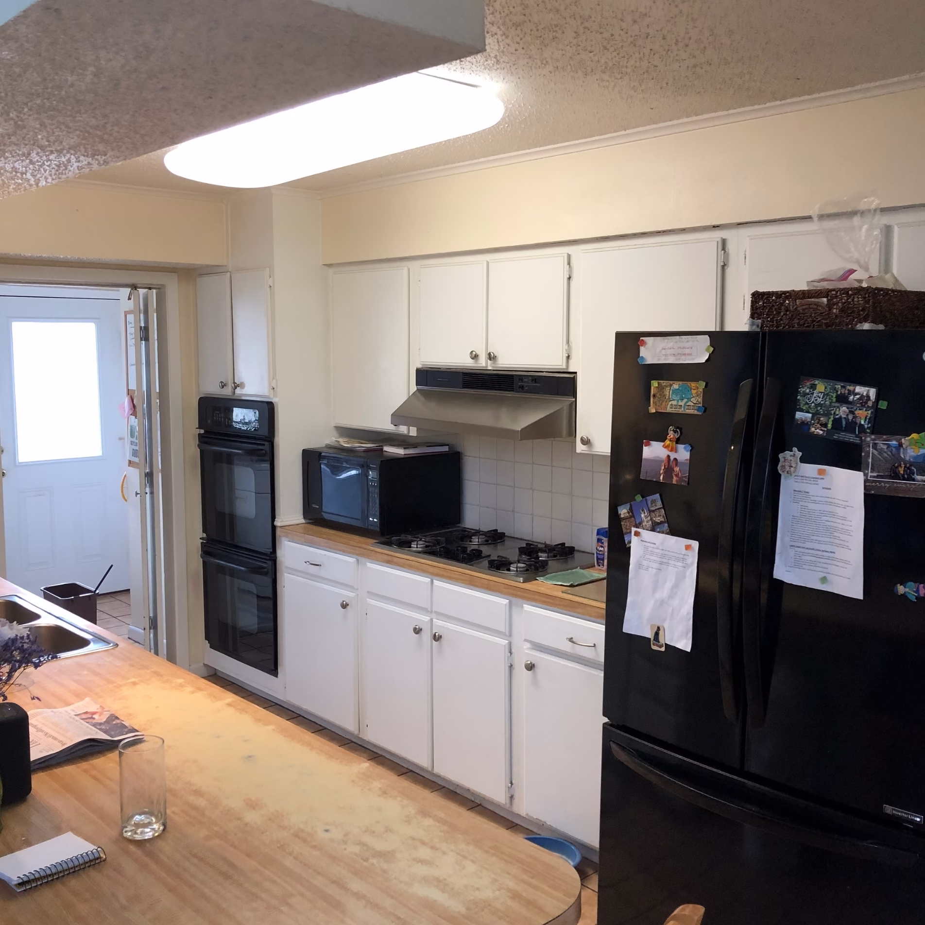 Cooking area before remodeling with white cabinetry, wood-look countertops, countertop microwave, gas cooktop, and original vent hood.