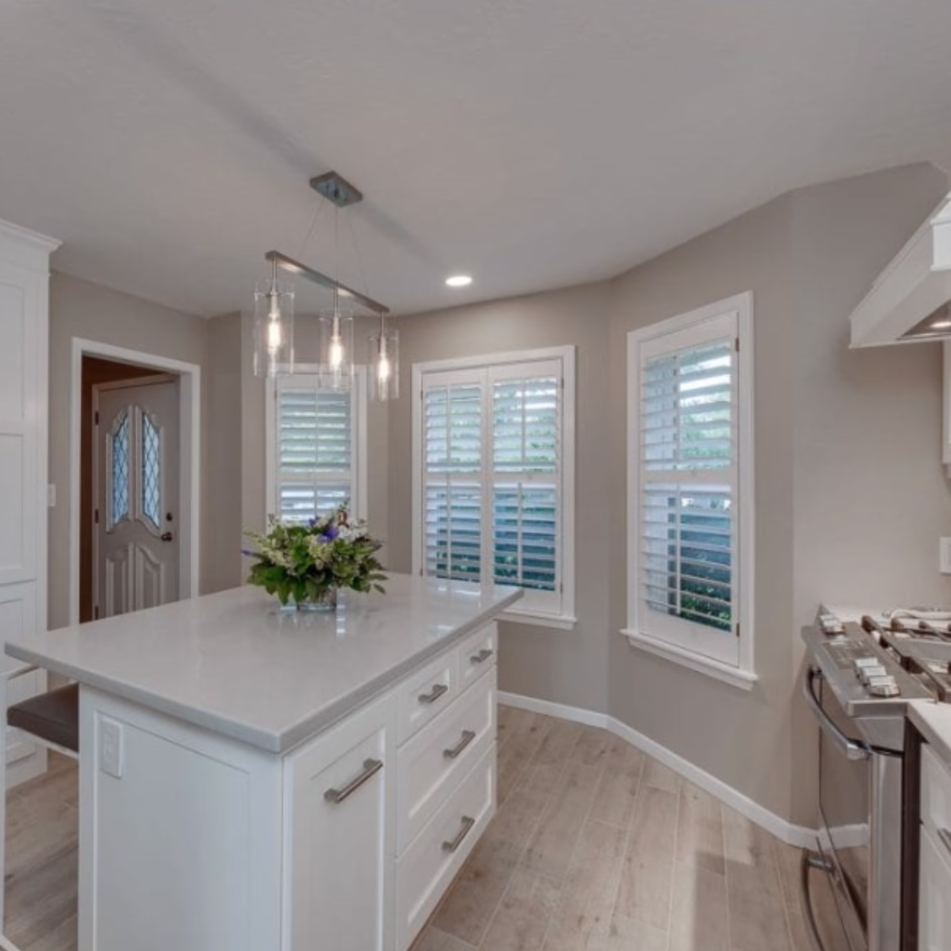 Remodeled kitchen with a white island, pendant lighting, wood-look flooring, and plantation shutters across three bay windows.