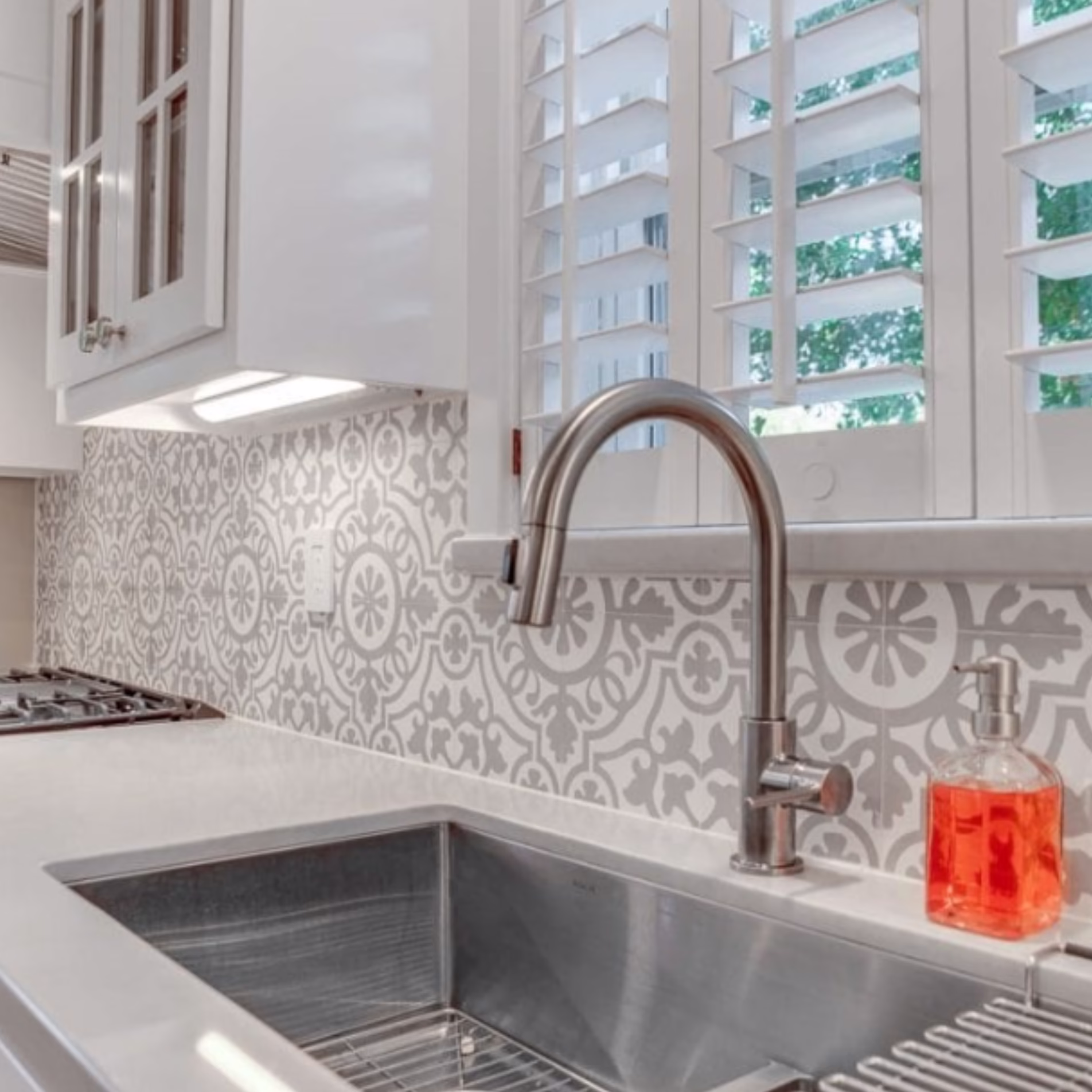Remodeled kitchen sink area with a stainless steel undermount sink, pull-down faucet, gray patterned backsplash, and white cabinetry.