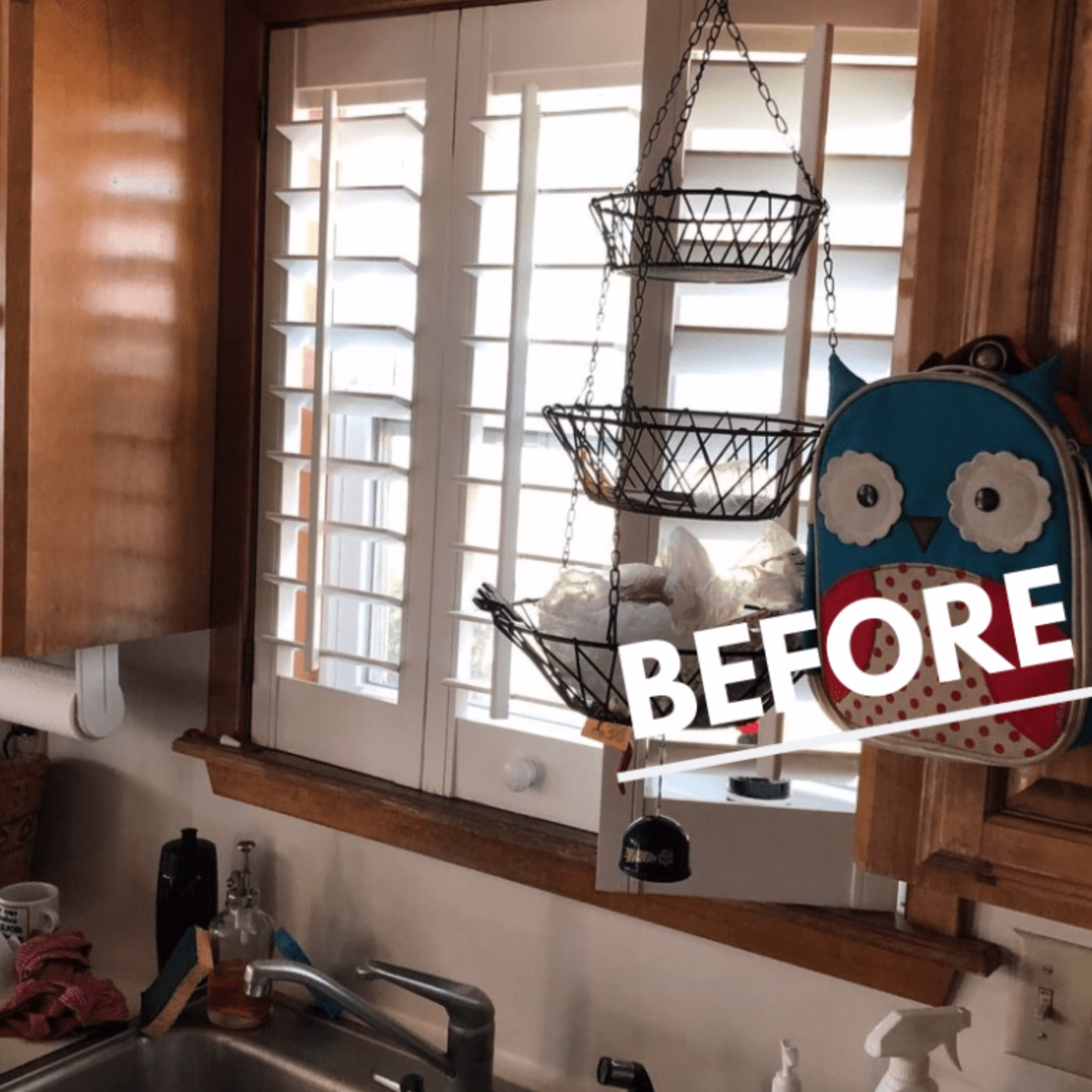 Original kitchen sink beneath plantation shutters, surrounded by natural wood cabinets, wood window trim, and hanging wire storage baskets.