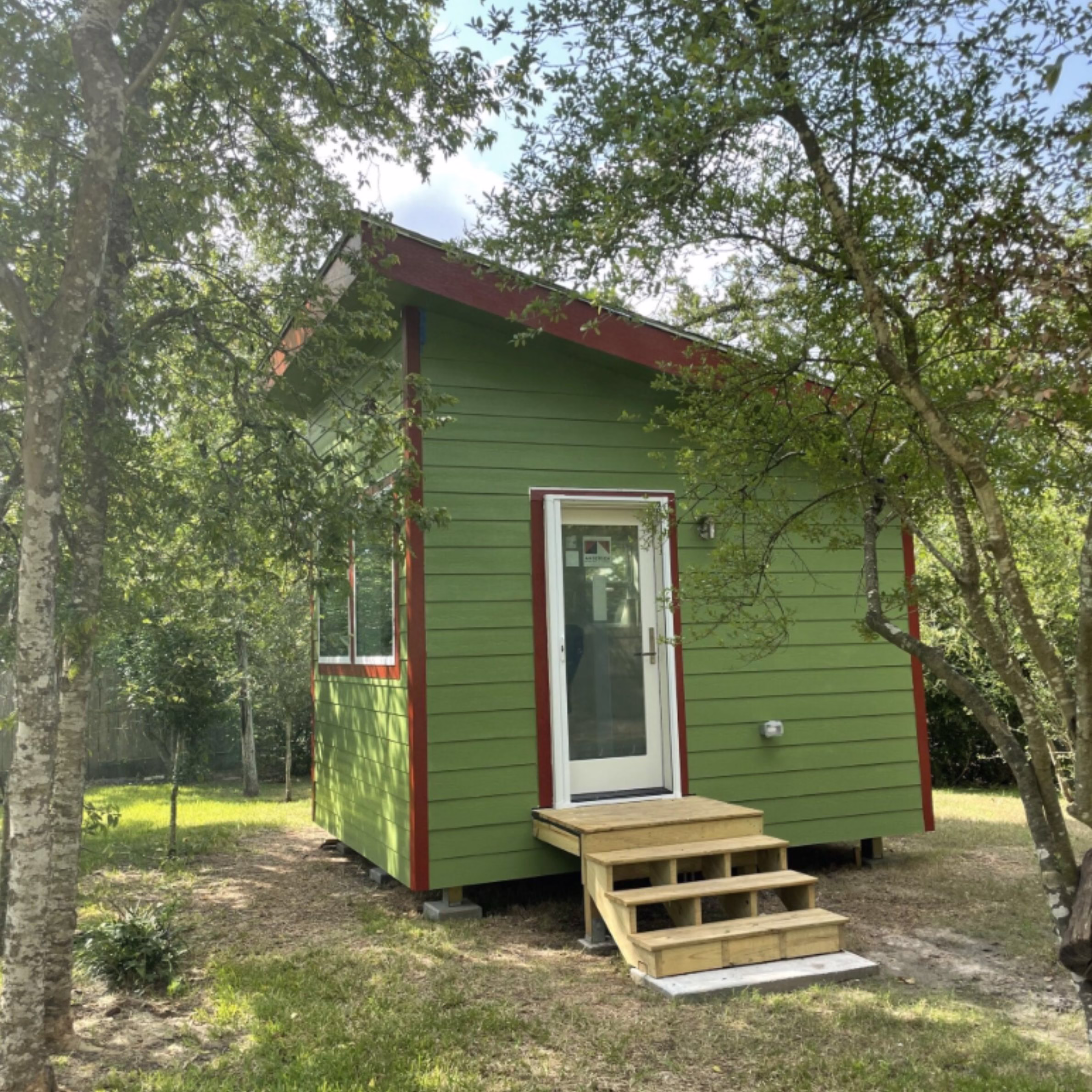 Completed green detached home office with red trim, entry steps, and a sloped roof surrounded by mature trees.