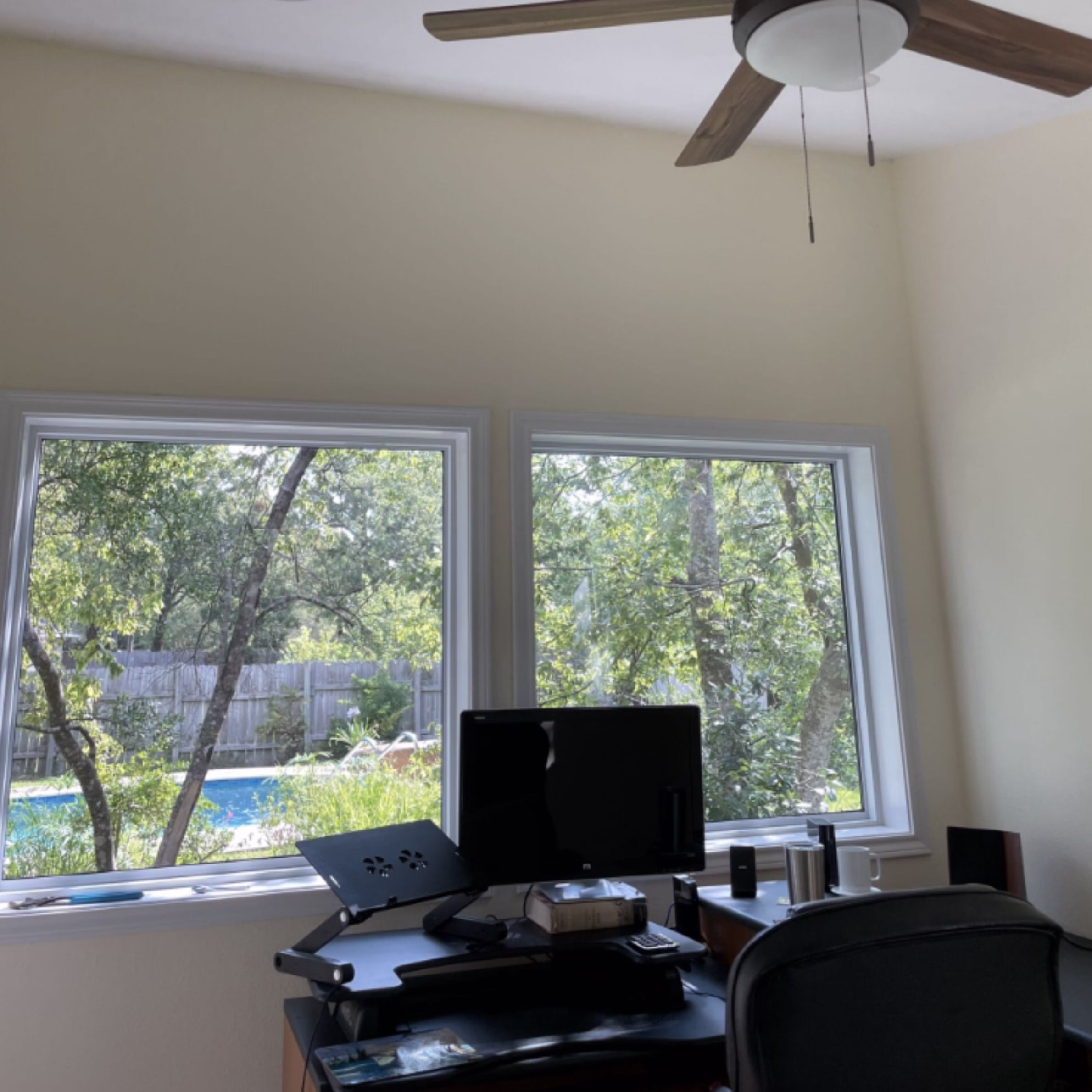 Finished home office interior with desk, computer monitors, ceiling fan, and large windows overlooking the backyard swimming pool.