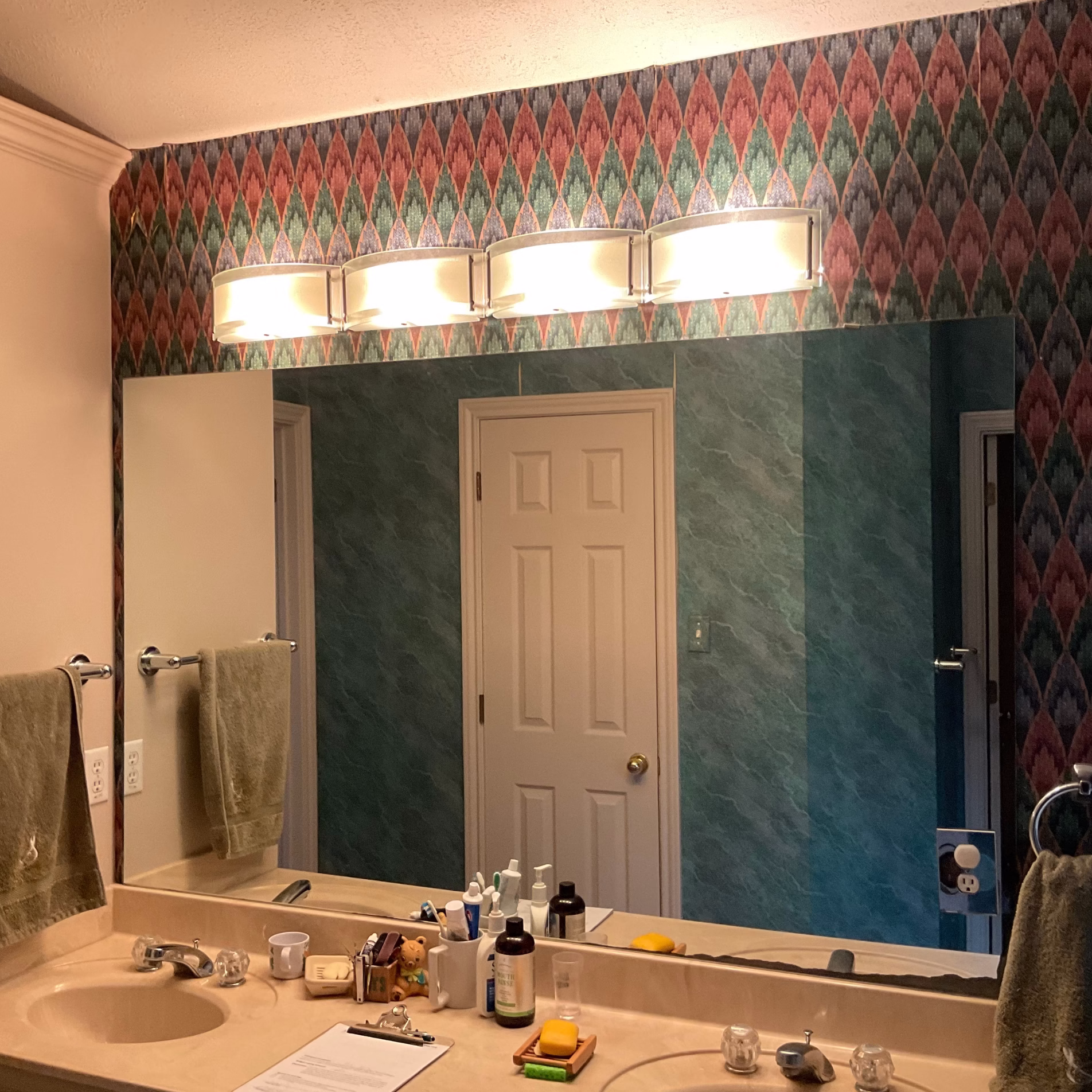 Primary bathroom vanity before remodeling with beige countertop, large wall mirror, patterned wallpaper, and original light fixture