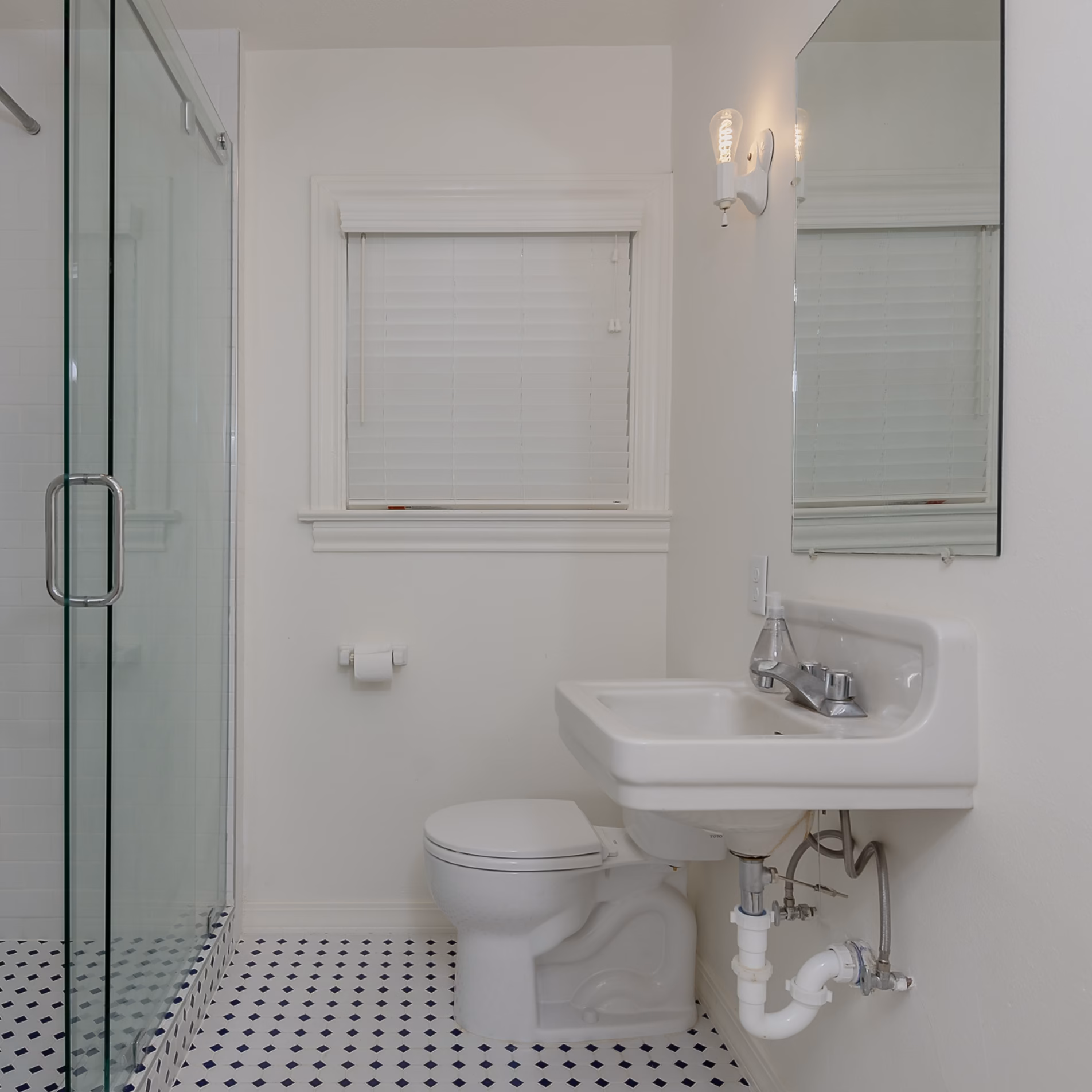 Remodeled guest bathroom with original white wall-mounted sink, glass-enclosed shower, and blue-and-white patterned tile flooring.