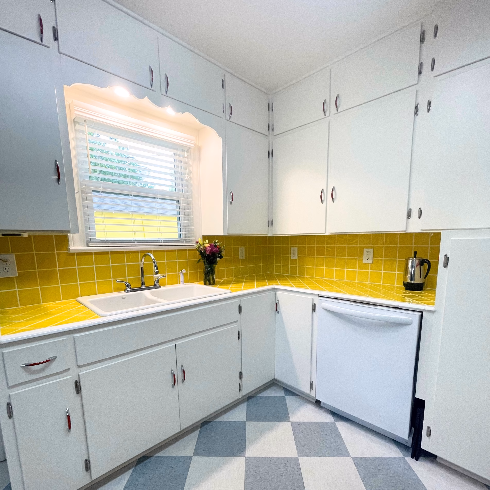 Remodeled 1950s kitchen in Bryan, Texas, featuring bright yellow tile countertops, white cabinets, red-accented handles, and checkerboard flooring.