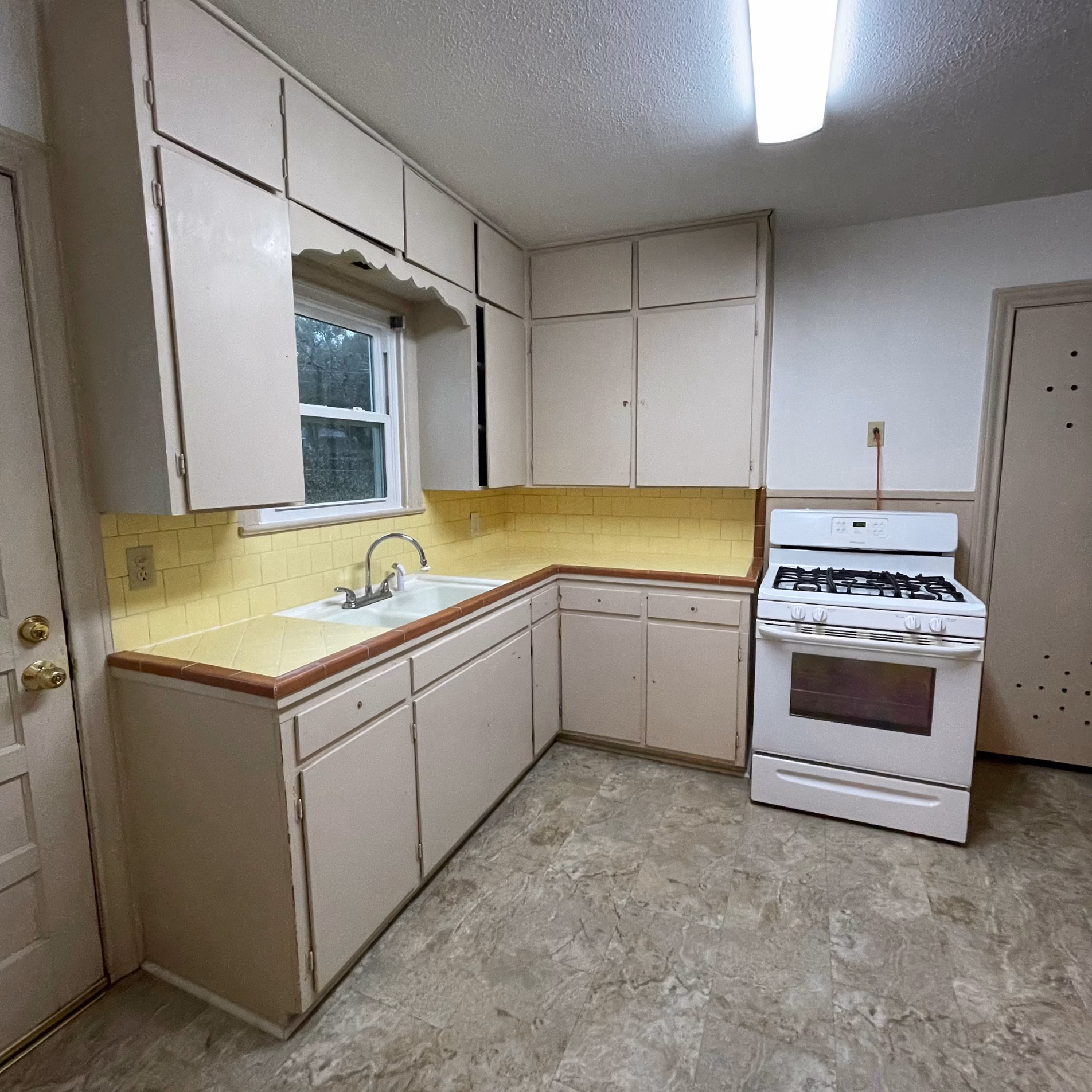 Original 1950s kitchen before remodeling, with pale yellow tile countertops, white cabinets, freestanding stove, and gray vinyl flooring.
