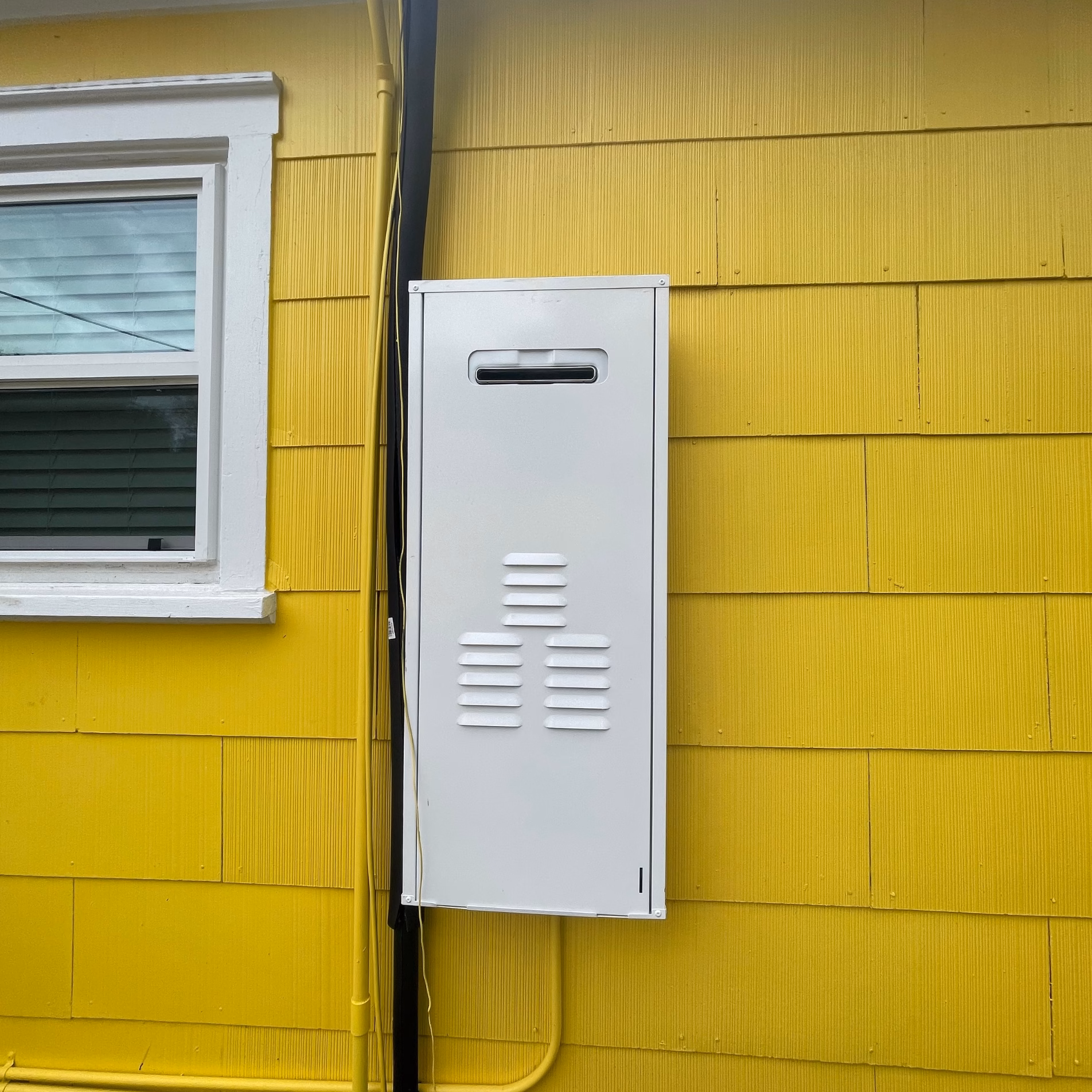 Exterior-mounted tankless water heater installed on the yellow siding of a remodeled 1950s home in Bryan, Texas.