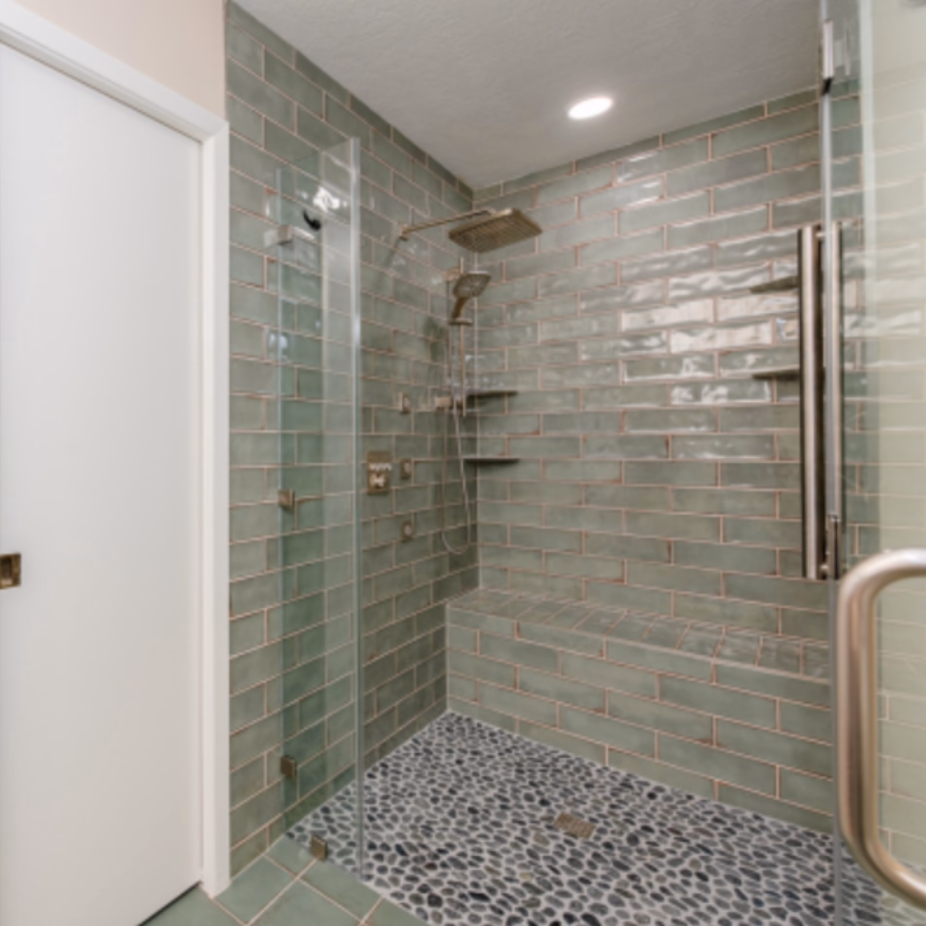 Remodeled curbless shower with green subway tile walls, black pebble floor tiles, built-in bench, and glass enclosure.