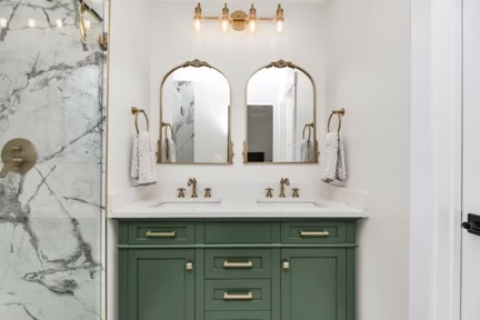 Remodeled bathroom with green cabinetry, dual arched mirrors, brass fixtures, and marble-patterned tile.