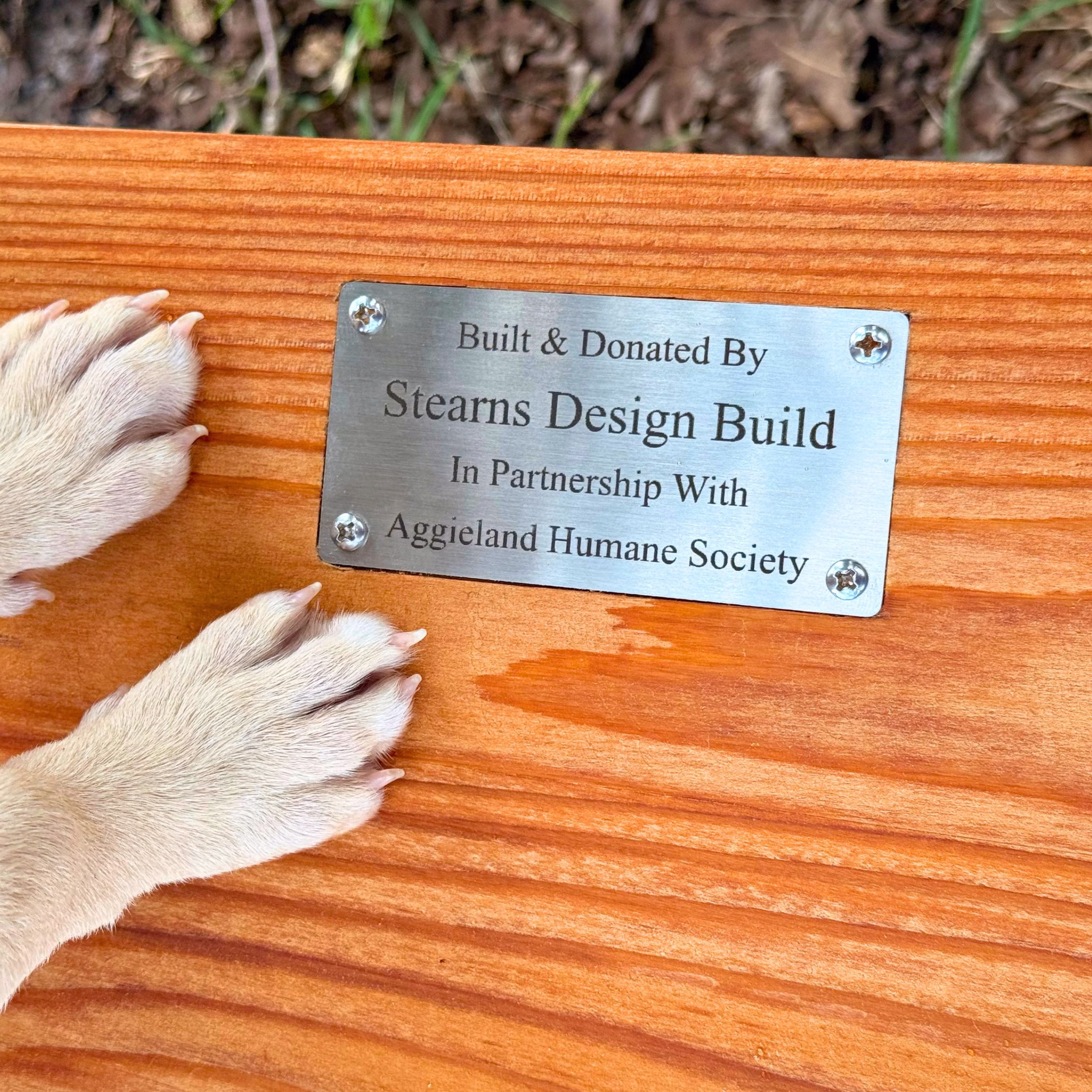 Custom cedar bench donated by Stearns Design Build to Aggieland Humane Society with puppy paws resting beside the partnership plaque in Bryan-College Station.