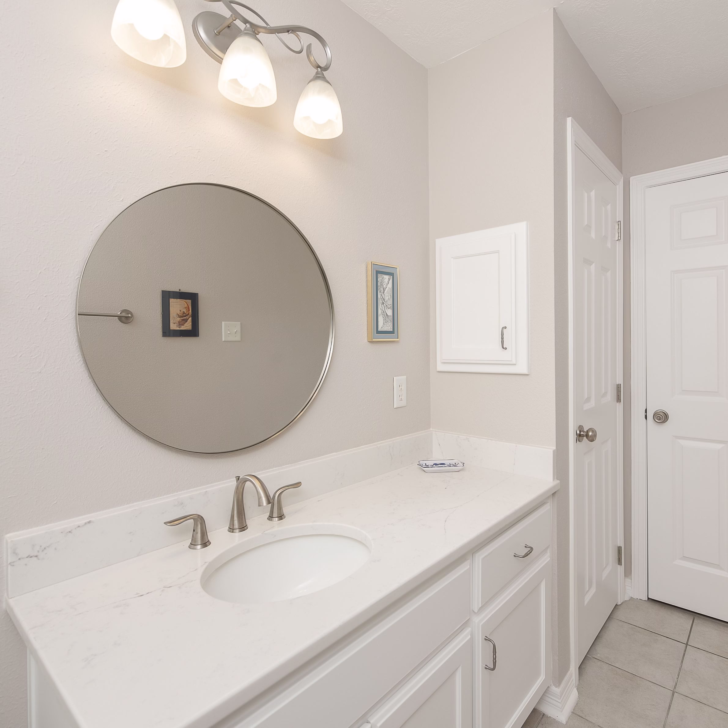 Modernized guest bathroom vanity completed during an aging in place whole home remodel, featuring quartz countertops, round mirror, updated lighting, and painted cabinetry.