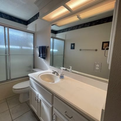 Guest bathroom vanity before an aging in place whole home remodel featuring original cultured marble countertop, large mirror, fluorescent lighting, and enclosed shower layout.