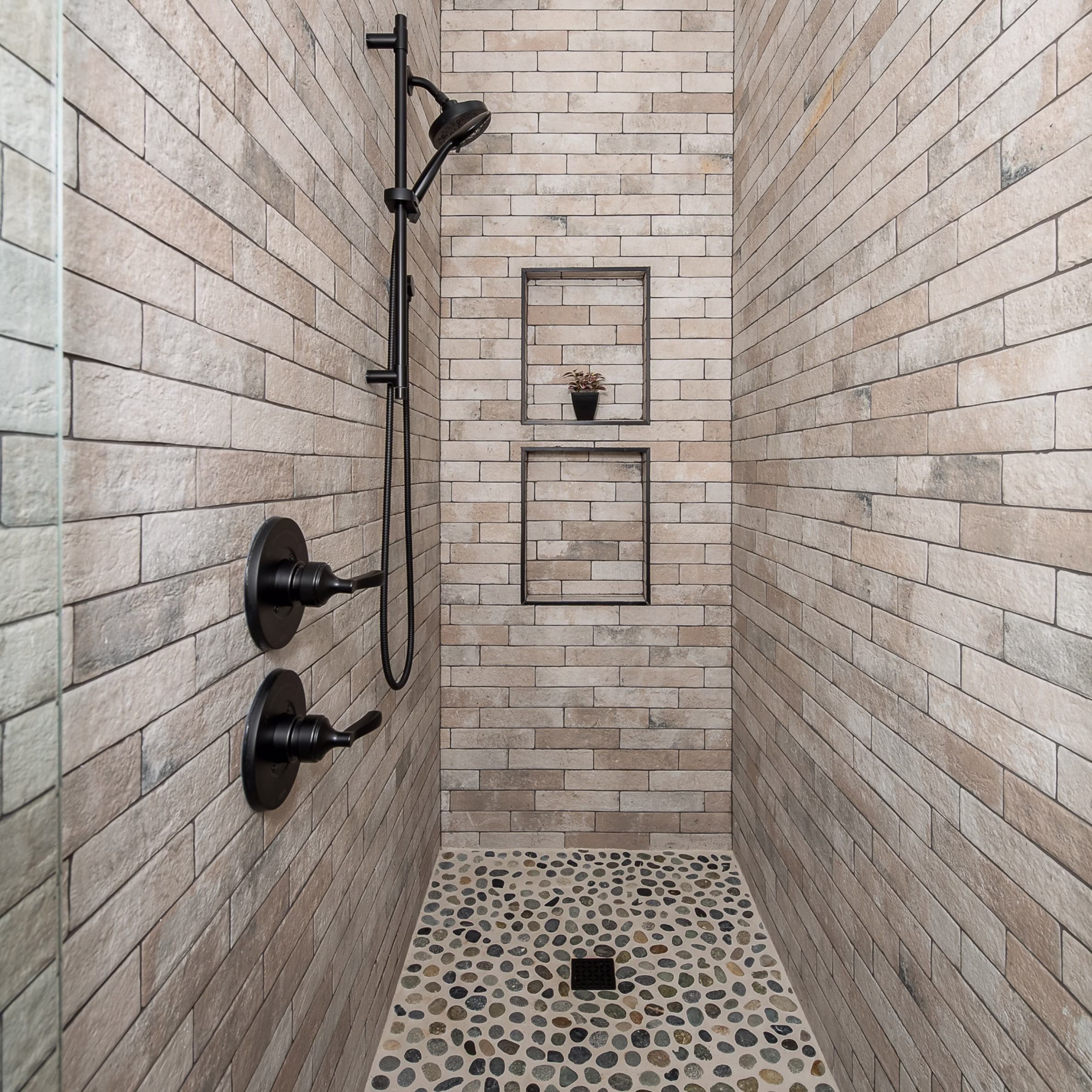 Narrow walk-in guest bathroom shower with full-height rectangular tile, pebble floor, black fixtures, and two recessed niches.
