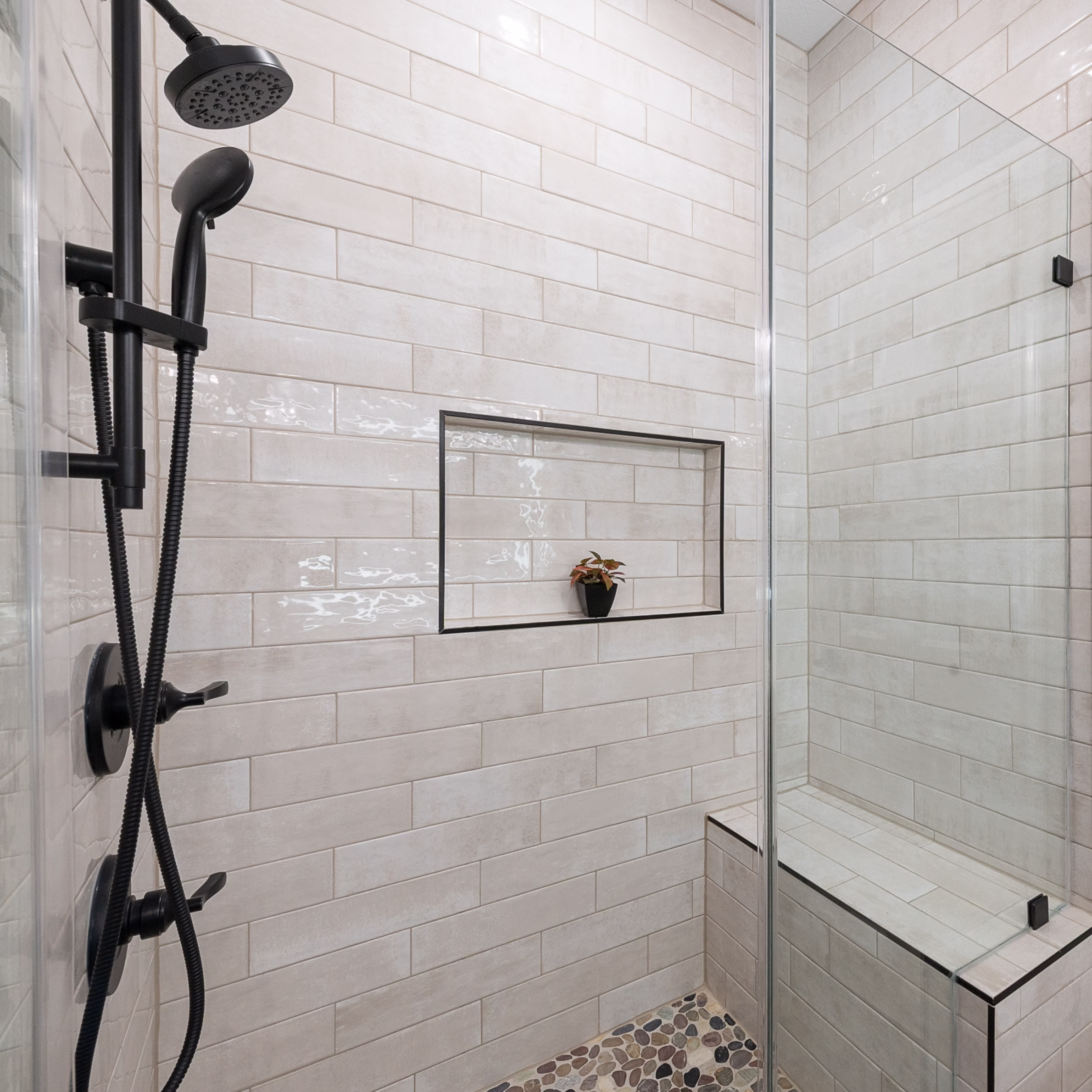 Glass-enclosed guest bathroom shower with light rectangular wall tile, pebble floor, built-in tiled bench, niche, and matte black fixtures.