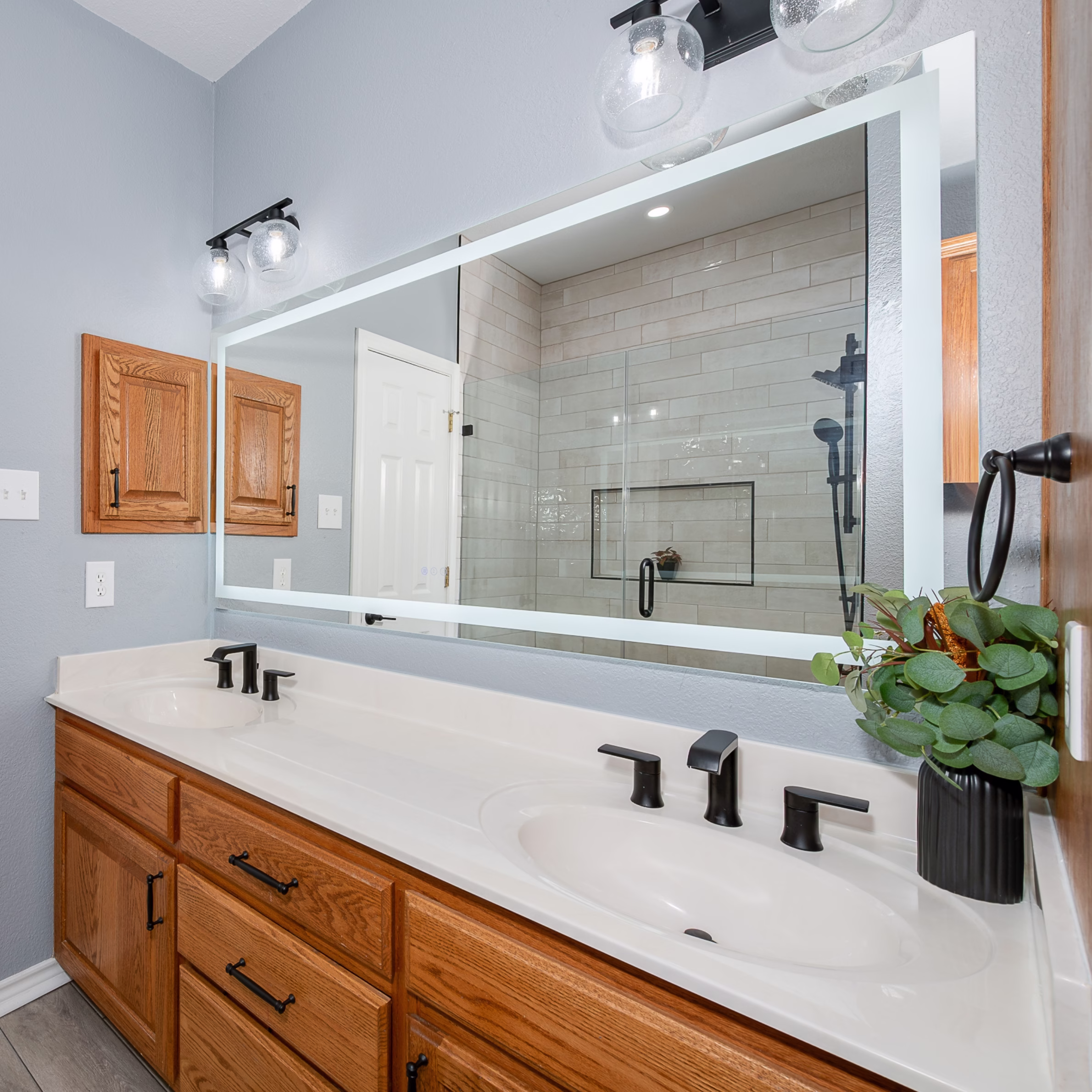 Double oak vanity with matte black faucets, illuminated mirror, and updated lighting in a remodeled guest bathroom in Bryan, TX.