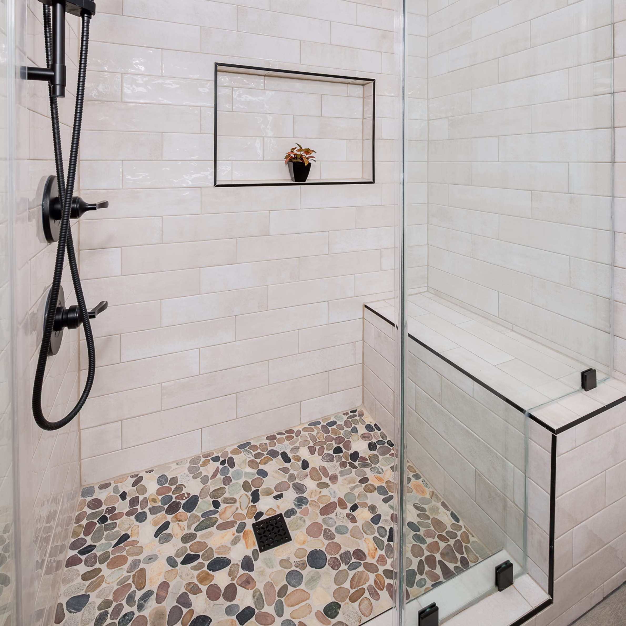 Tiled walk-in guest bathroom shower with glass enclosure, built-in bench, wall niche, pebble shower floor, and matte black fixtures.