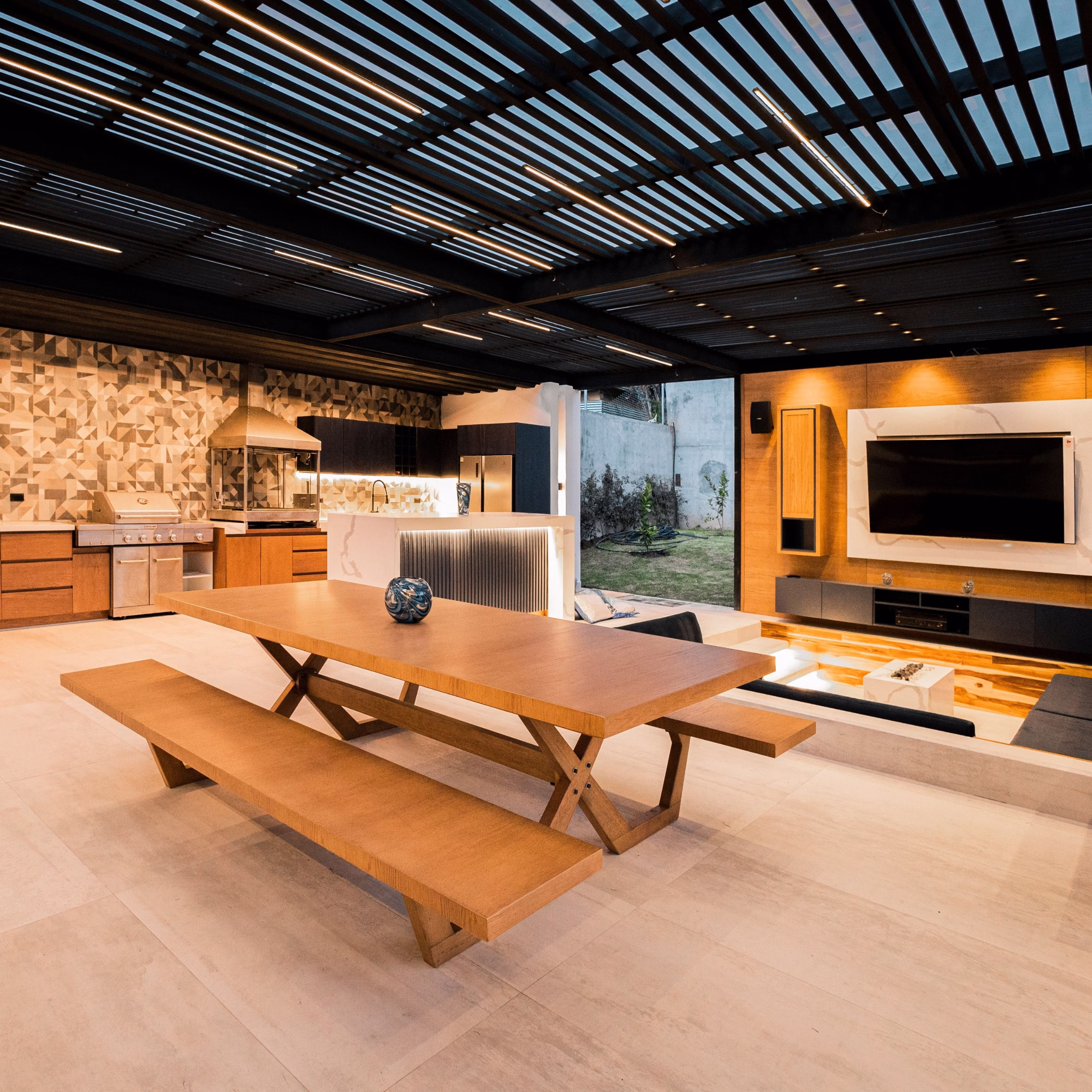Large covered outdoor kitchen and entertaining space with grill, sink, refrigerator, dining table, lounge area, and integrated lighting.