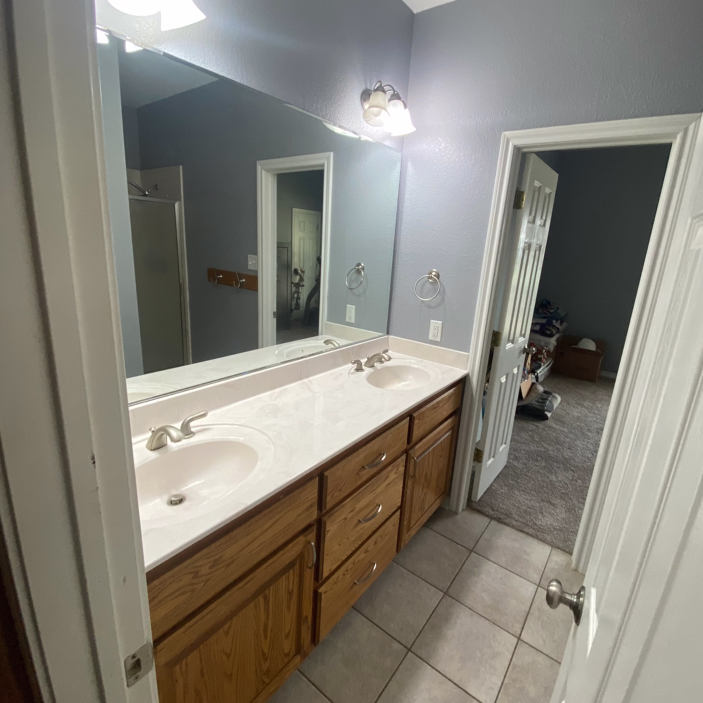 Guest bathroom before remodeling with an oak double vanity, white countertop, large wall mirror, and beige tile floor.