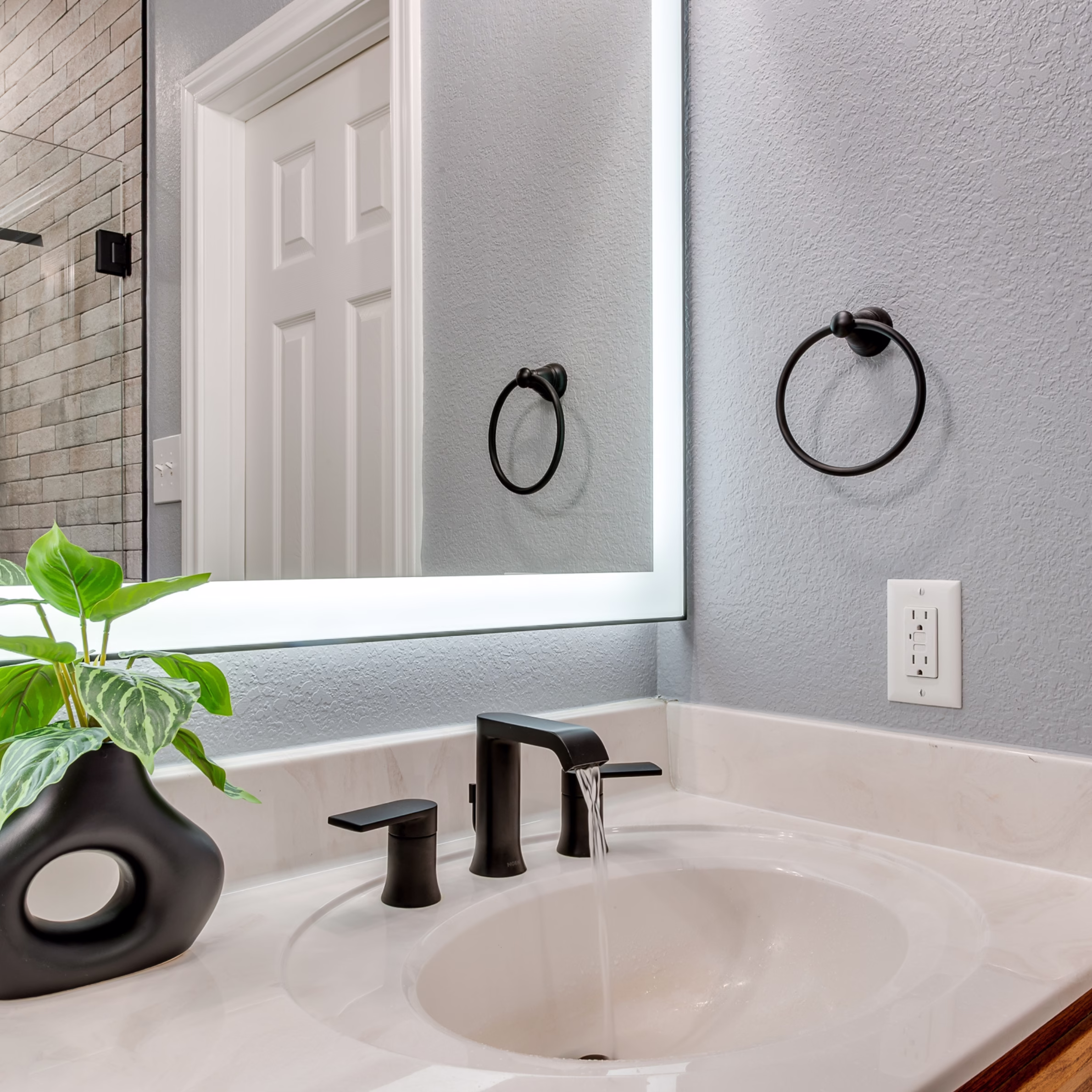 Guest bathroom vanity with matte black faucet, illuminated mirror, white countertop, and retained oak cabinetry.