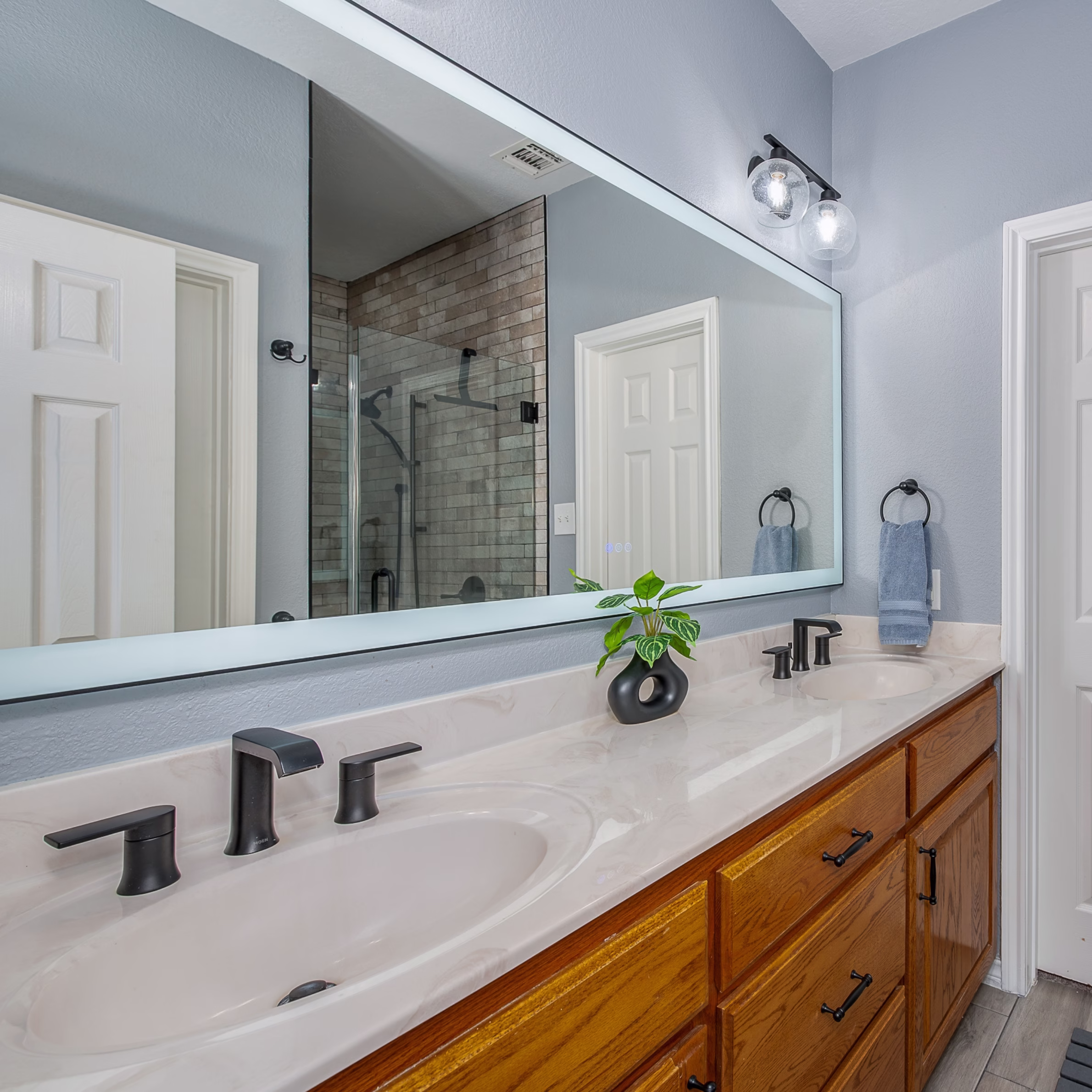 Retained oak double vanity updated with matte black hardware and faucets beneath a large illuminated mirror.