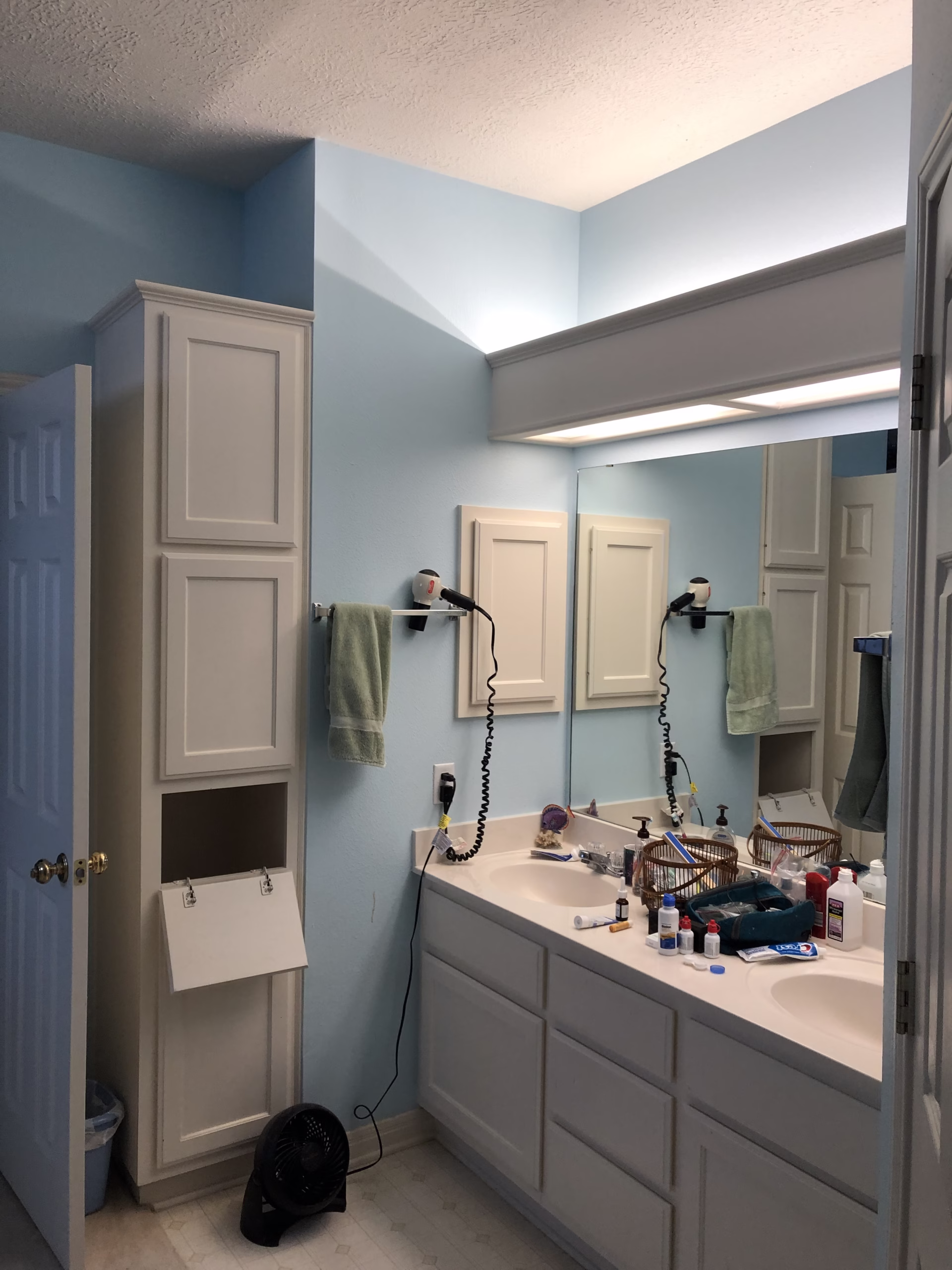 Original builder-grade bathroom with a double vanity, large wall mirror, overhead lighting and limited countertop storage.