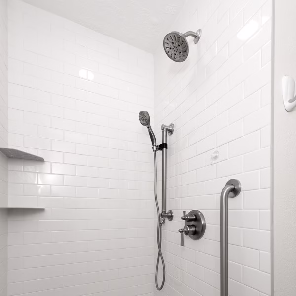 Curbless walk-in shower with white subway tile, grab bar, hand shower, and built-in corner shelves for improved accessibility.