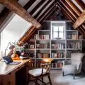 Attic home office with exposed wooden beams, a built-in bookshelf, a desk, and an upholstered armchair.