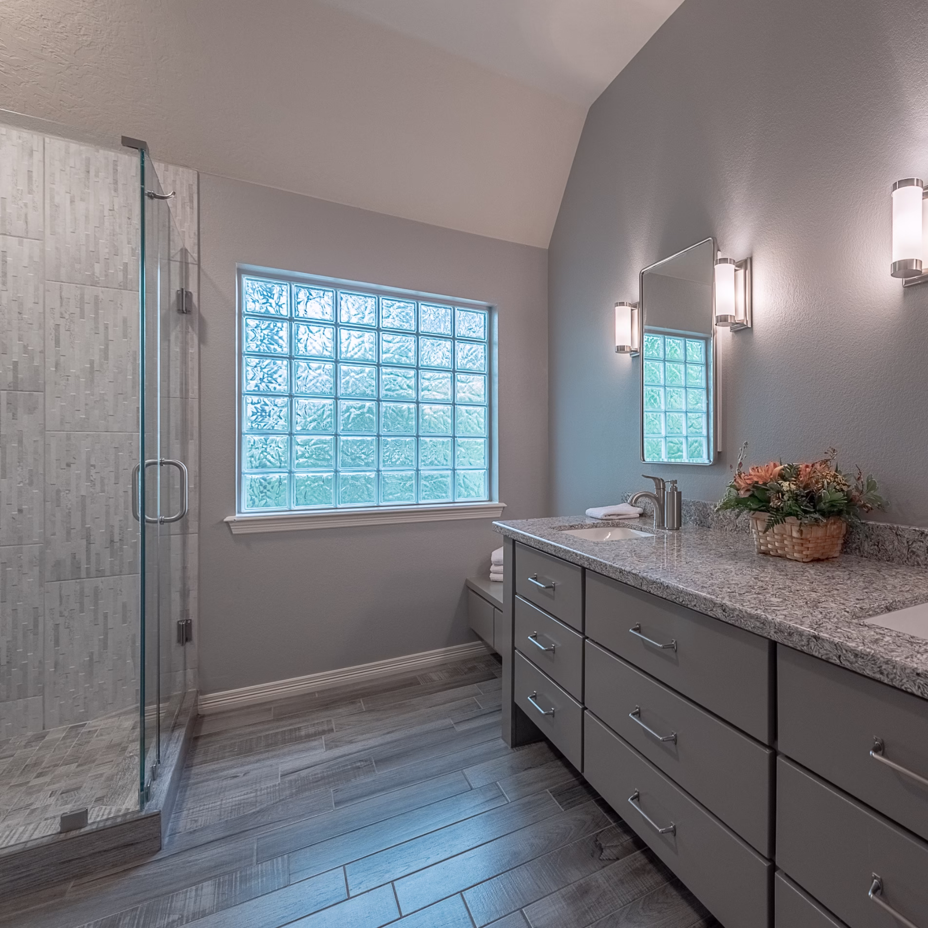 Finished primary bathroom with a frameless glass shower, glass block window, gray double vanity, and wood-look tile flooring.