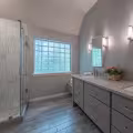 Finished primary bathroom with a frameless glass shower, glass block window, gray double vanity, and wood-look tile flooring.