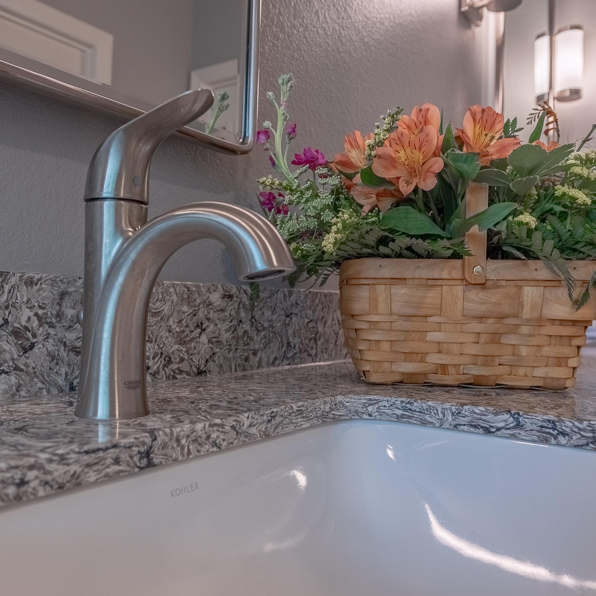 Close-up of the remodeled bathroom vanity with brushed metal faucet, undermount sink, stone countertop, mirror, and floral arrangement.