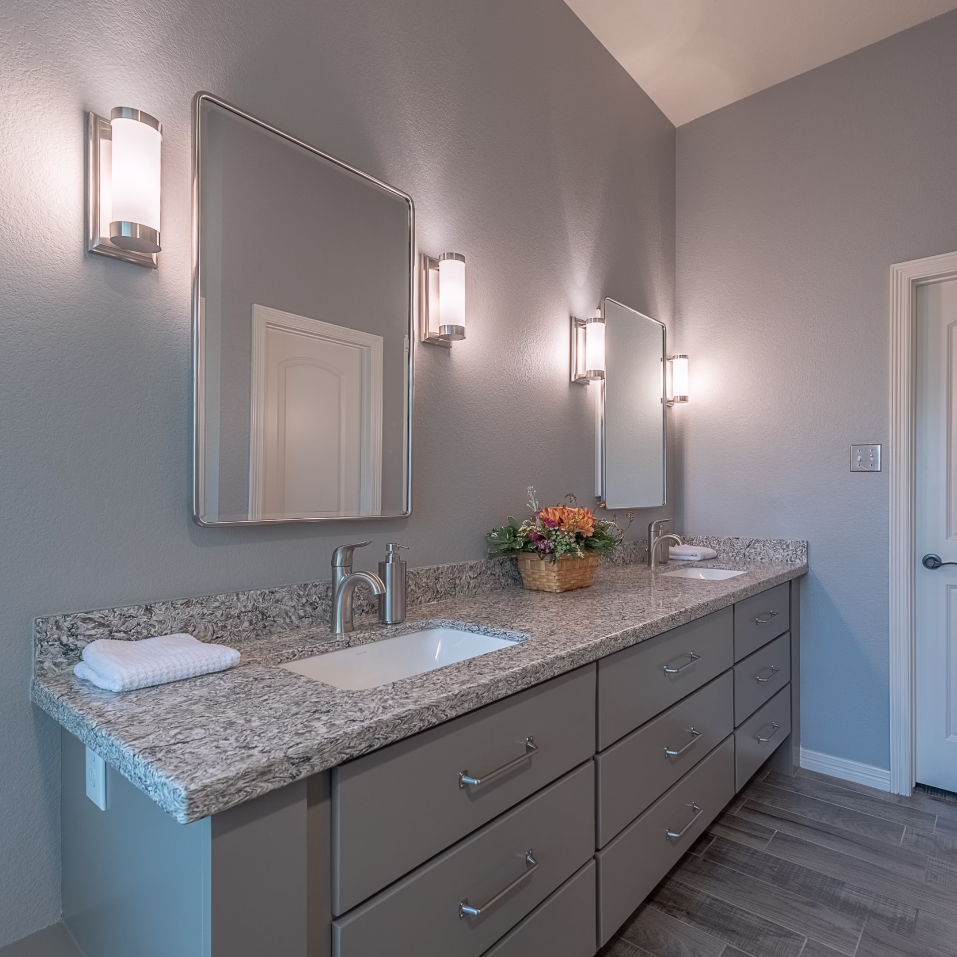 Remodeled double vanity with gray cabinetry, stone countertop, undermount sinks, individual mirrors, and wall sconces.