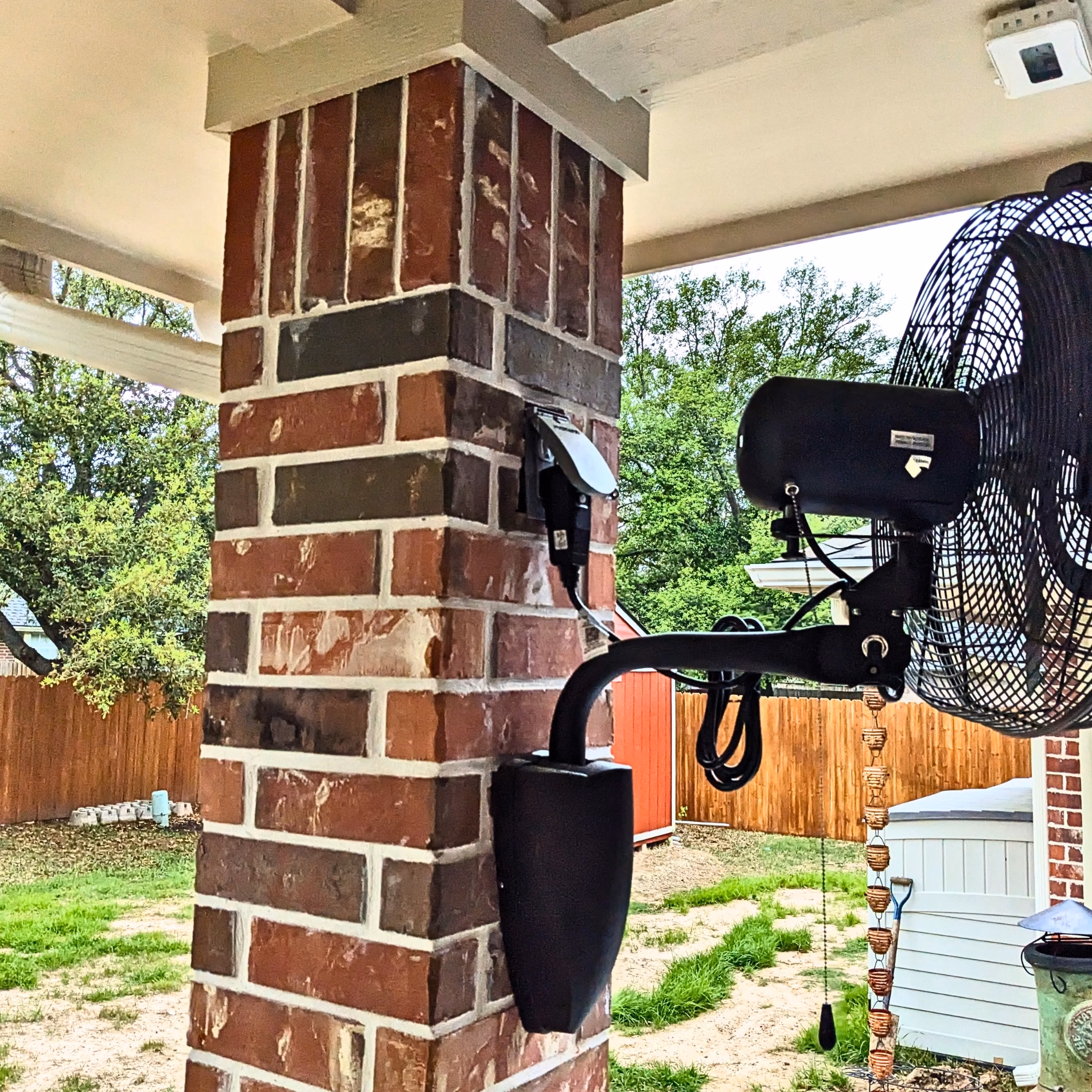 Black oscillating wall-mounted fan installed on a brick column beneath the expanded covered patio roof.