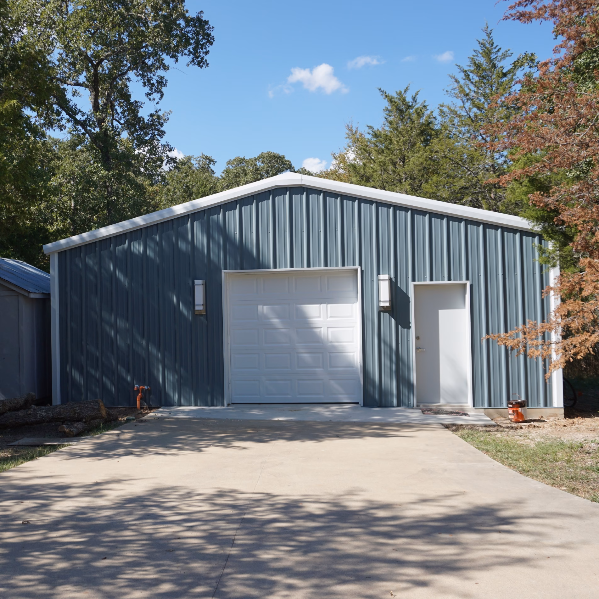 Newly constructed blue-gray metal barn surrounded by preserved mature trees, with tree shadows extending across the building and driveway.